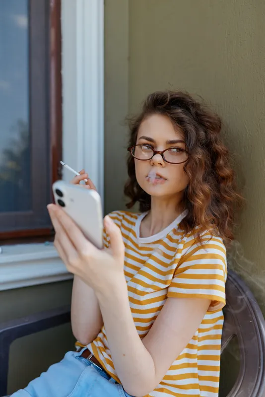 Casual woman on a sunlit outdoor porch holds a phone, wearing striped tshirt and glasses, capturing a candid lifestyle moment with relaxed contemporary vibe and warm daylight.