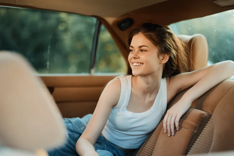 Young smiling woman in casual clothing enjoying a sunny day inside a car, embodying joy and relaxation with a natural background and warm tones.