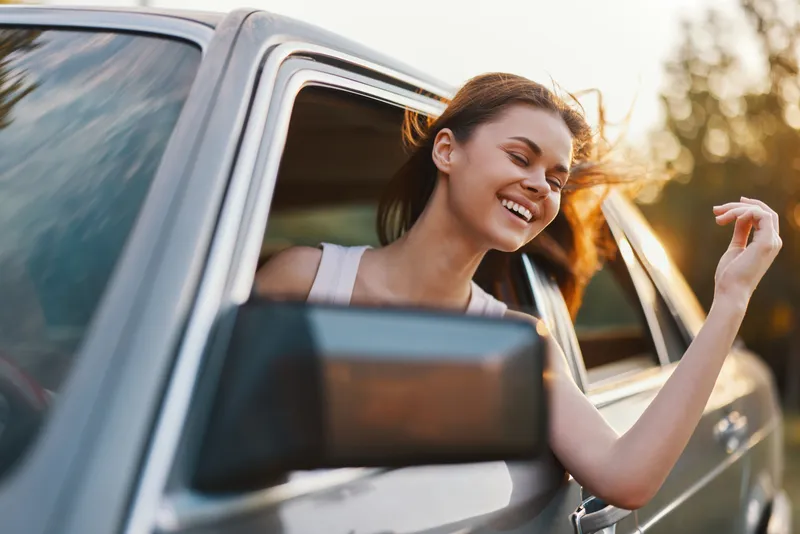 Happy young woman enjoying a road trip with a breeze in her hair, leaning out of a car window, capturing the essence of freedom and adventure during golden hour.