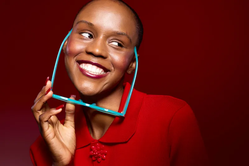 Portrait of a smiling black woman with short hair wearing a red sweater holding blue eyeglasses near her face. Bright studio lighting and red background create a vibrant cheerful mood.