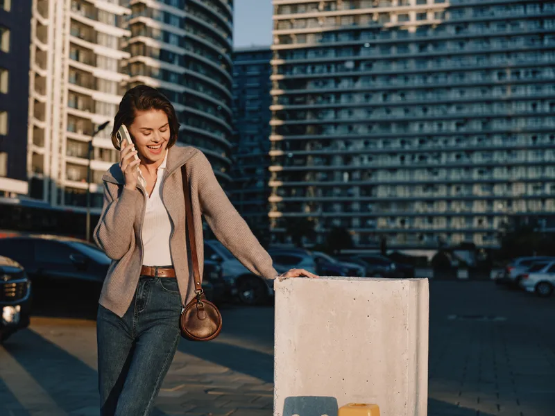 A cheerful woman walks outdoors in a modern city, talking on a smartphone. She smiles and appears engaged, creating a positive, energetic urban atmosphere.