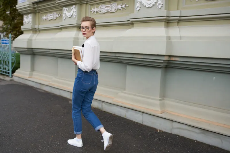 Молодая женщина в джинсах и рубашке с книгой в руках рядом с фасадом здания, образование