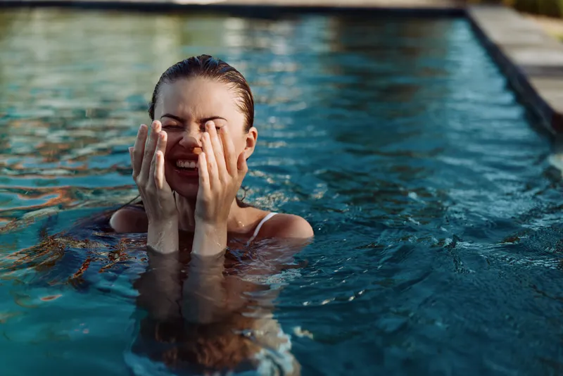 Woman in a swimming pool covers her face with hands, expressing surprise and relief, a candid lifestyle moment with water, sun, and warm mood, soft ripples enhance calm