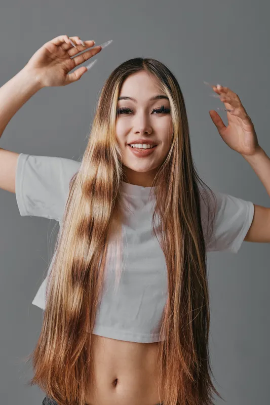 Young woman smiling with long straight hair wearing casual white t shirt posing with raised hands on gray background. Beauty, lifestyle and hair care concept with natural light.