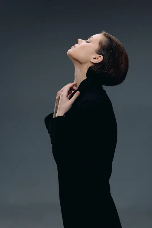 Profile of a woman in black clothing with eyes closed, expressing serenity and elegance against a dark gray backdrop.