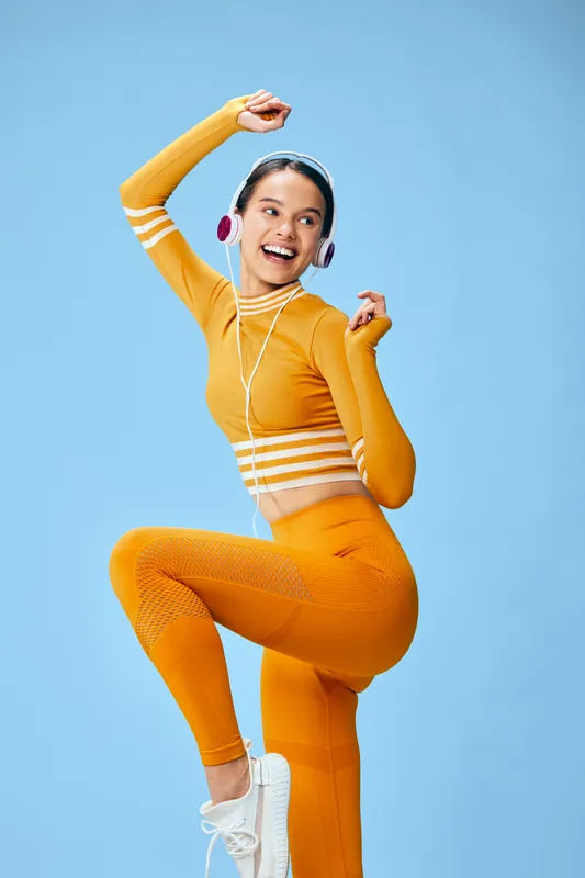 Dynamic female athlete in orange activewear dances midair against a blue studio backdrop, wearing headphones and a bright smile, showcasing energy, movement and a vibrant fitness lifestyle.