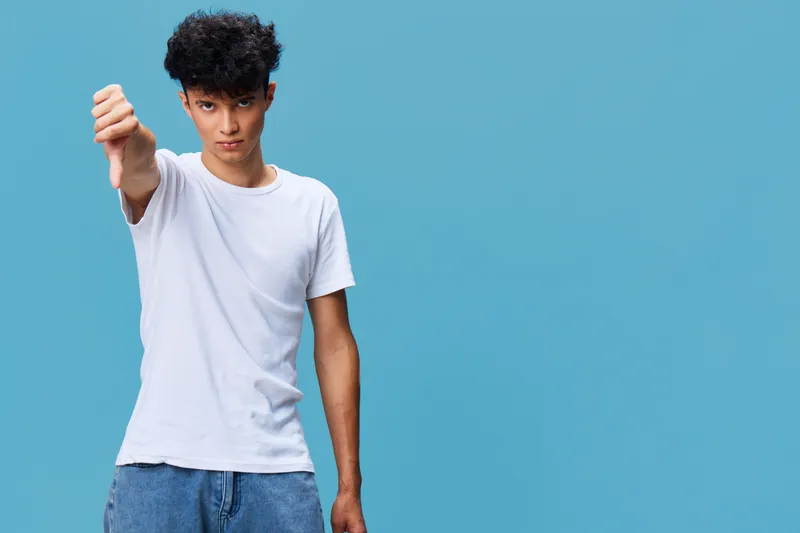 young man in white t-shirt making thumbs down gesture with displeased expression on blue background, casual style, negative emotion, isolated