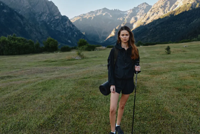 A young woman stands alone in a wide meadow framed by towering mountains. She wears a dark jacket and shorts, holding a hiking pole and hat, looking confident and calm as nature surrounds her.
