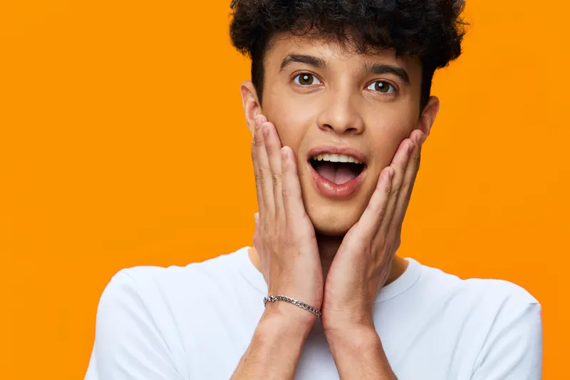 surprised young man with curly hair wearing white shirt on vibrant orange background expressing shock and amazement with open mouth and hands on cheeks.