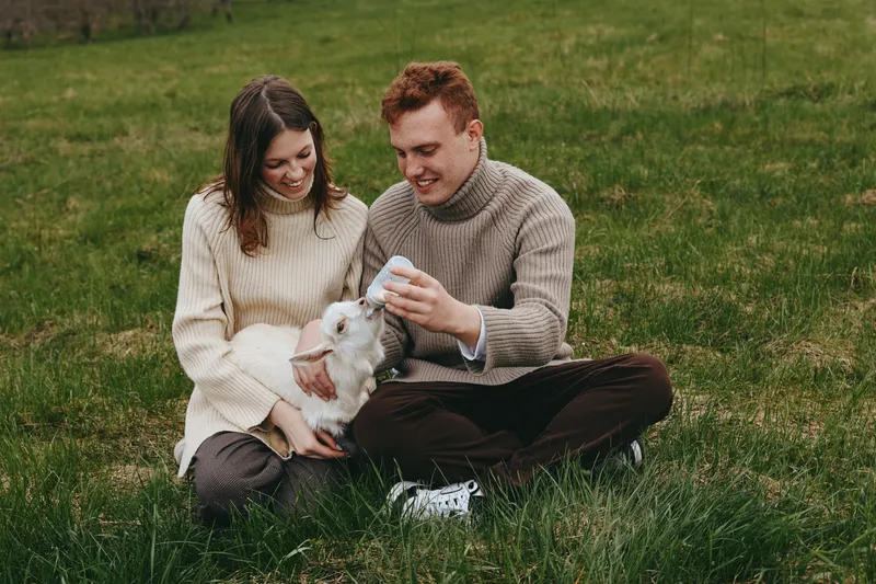 Couple relaxing in a grassy field gently interacting with a playful goat kid, showcasing affection and outdoors lifestyle, warm natural lighting, candid wholesome moment.