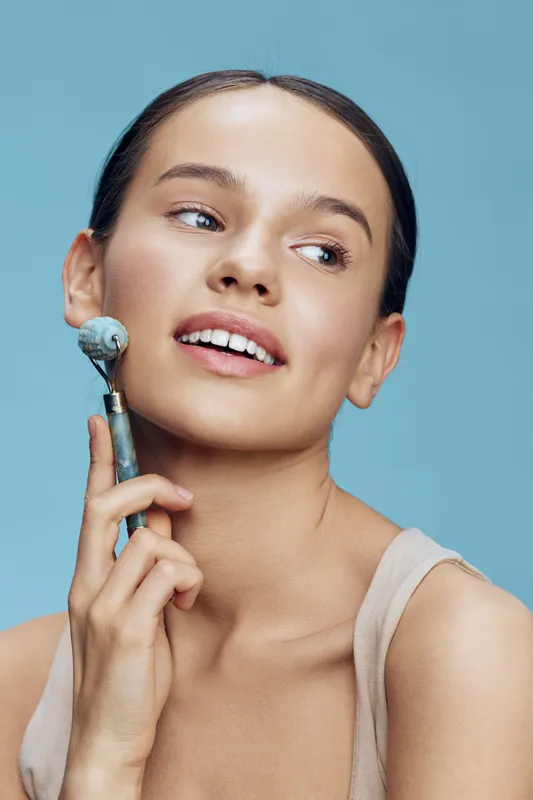 Young woman with smooth complexion using a jade roller on her face, showcasing a fresh, radiant look against a light blue background