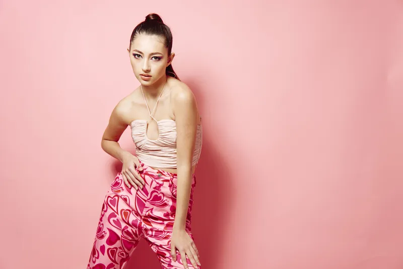 Young woman poses pursuing fashion trends in a stylish strapless top and colorful pants with heart patterns against a pink background. Beauty concept with creative vibes.