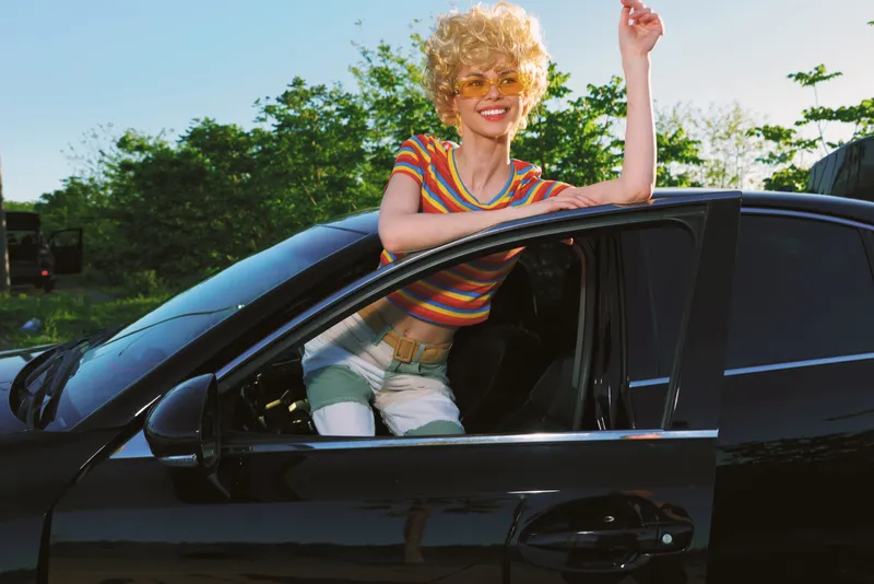Young woman with curly blonde hair leans out of a black car window, smiling and raising her arm in a carefree summer moment, wearing a striped tee and light shorts with sunlit greenery in the