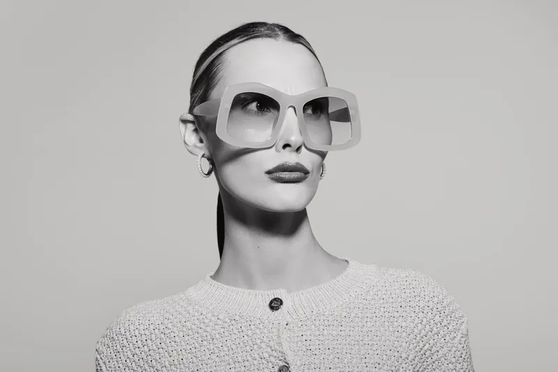 Stylish white woman with pale skin wears oversized sunglasses and knitted top. Studio portrait with elegant makeup and slicked back hair in monochrome. Concept of fashion and confidence.
