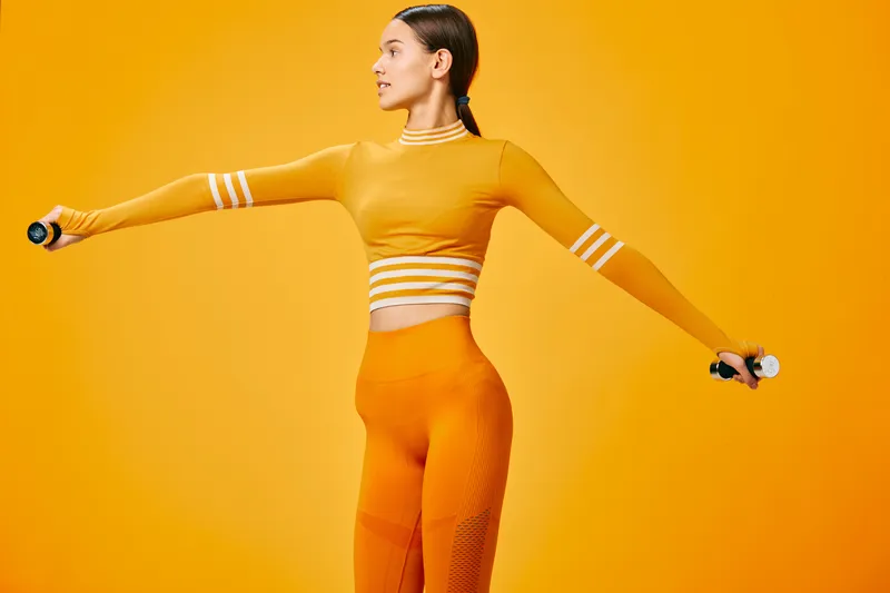 Young woman fitness athlete exercising with small dumbbells in matching orange activewear, arms extended for lateral raises against a bright monochrome orange studio background.