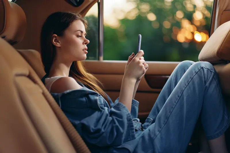Young woman using smartphone in the car during sunset, dressed in casual denim outfit, conveying relaxation and tranquility.
