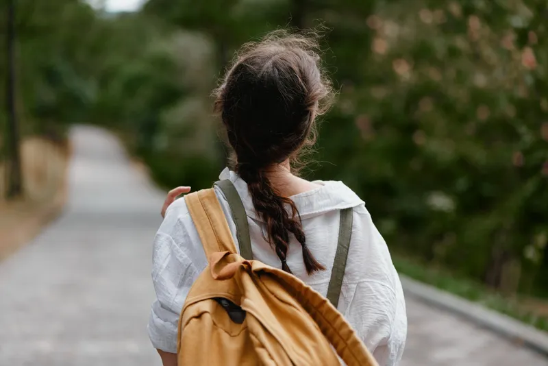 Young woman walking in nature with a backpack, enjoying serenity and a calm atmosphere, dressed casually in a white shirt, with a braided hairstyle and surrounded by greenery