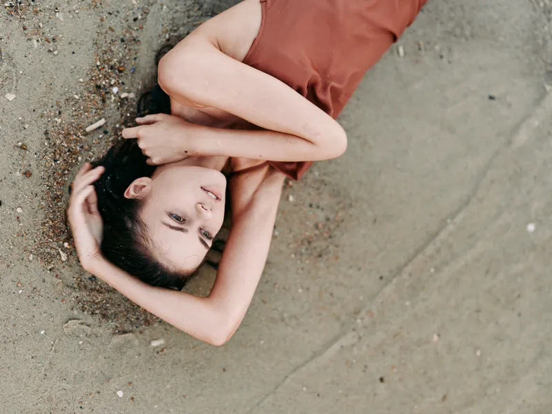 A young woman lies on sandy beach, eyes closed, arms raised over her head, wearing a rust sleeveless dress. The calm composition highlights relaxed body lines against grainy sand.