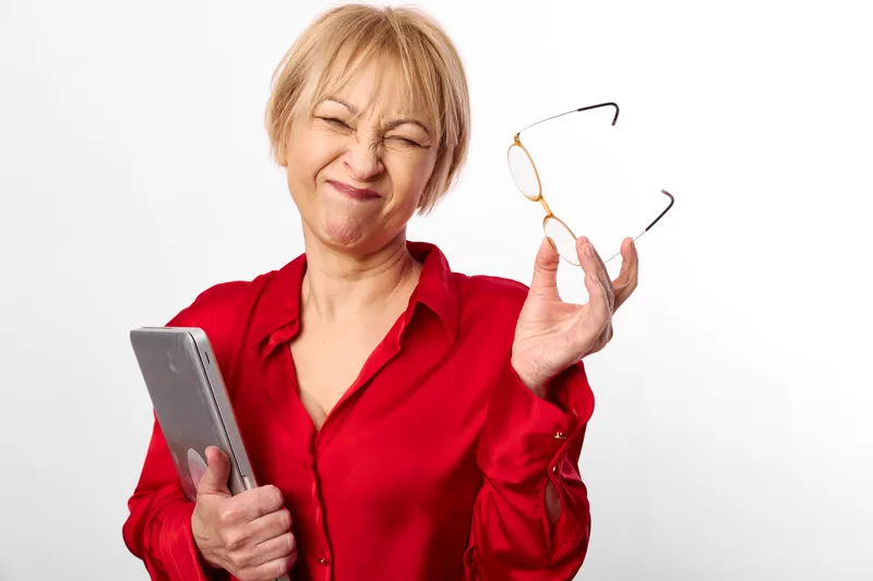 Senior woman in red blouse, holding a tablet, smiling while adjusting glasses, studio portrait on white background, daily technology concept, independence and friendly mood