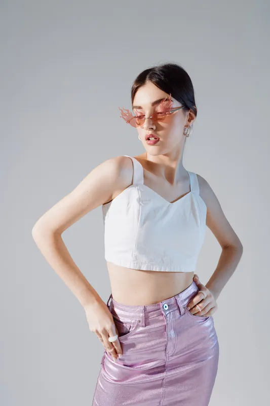 Young woman in pink sunglasses wearing a white crop top and shiny skirt poses confidently against a plain background. Concept of beauty fashion and style.