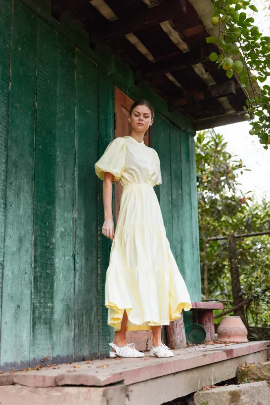 A woman wearing a yellow dress stands on a wooden porch beside a green wall, outdoors in a garden setting, exuding style and calm confidence and grace.