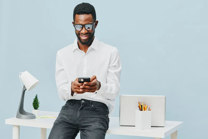 Smiling man using mobile phone in bright modern office workspace with laptop and desk accessories, casual outfit, cheerful mood, light blue background, lifestyle concept.