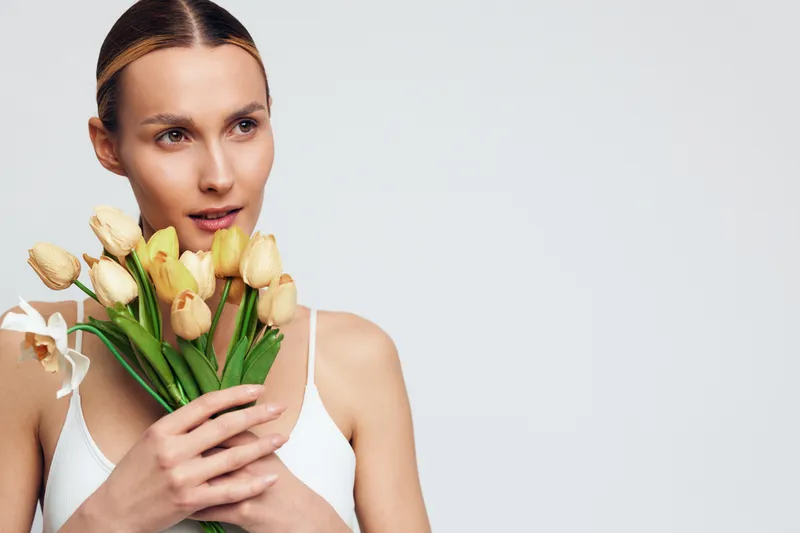 Young woman holding yellow tulips close to her face, thoughtful expression, soft lighting, minimalistic background, natural beauty, spring flowers, calm mood