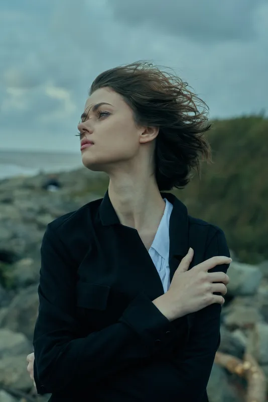 Wind swept coast, woman in dark jacket gazes away from the sea, wind teases her hair, moody mood, outdoor scene, nature and weather, evocative and dramatic