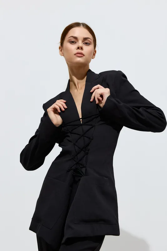 Fashion woman in black lace-up jacket with a serious expression posing against a plain white background, showcasing modern style and minimalist elegance.