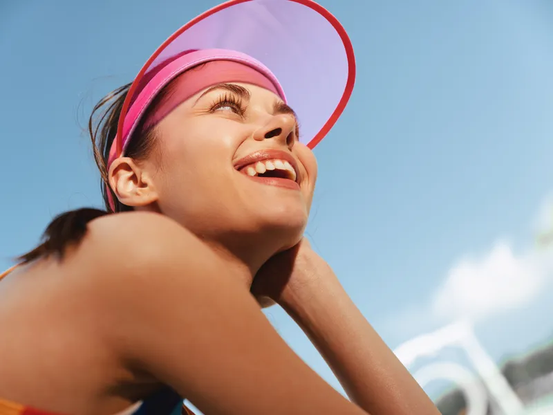 Bright portrait of a smiling girl wearing a pink visor, looking upward with joy under a clear blue sky, capturing playful summer energy and carefree outdoor youth.