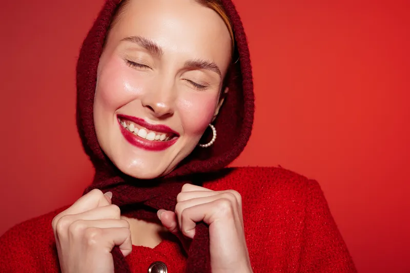 Smiling woman with closed eyes wearing red knitted scarf and sweater on red background. Concept of warmth, happiness, winter fashion, and cozy lifestyle with joyful expression.