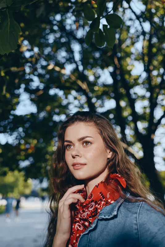 young woman with red scarf and denim jacket standing outdoors in sunny park with green trees and soft natural light, thoughtful expression
