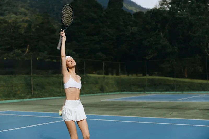 Young female athlete celebrating a tennis victory on a sunny court, showcasing joy and confidence in sporty attire against a lush green background