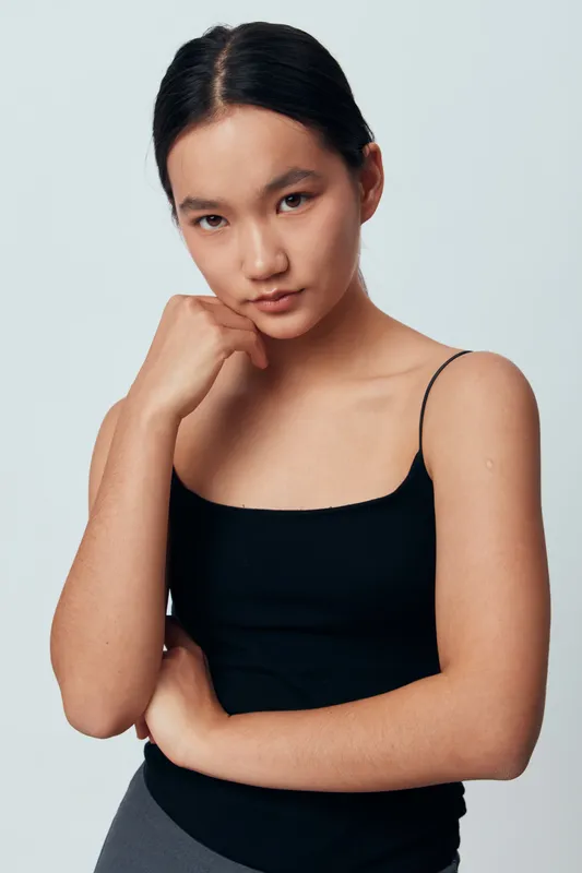 Young woman with natural makeup and dark hair wearing black tank top posing with hand near chin on light background for portrait and beauty concept.
