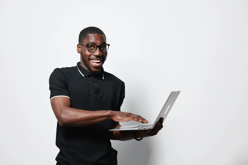 African American man with dark skin and glasses smiling while holding laptop and typing on keyboard isolated on white background. Casual black polo shirt and positive expression.