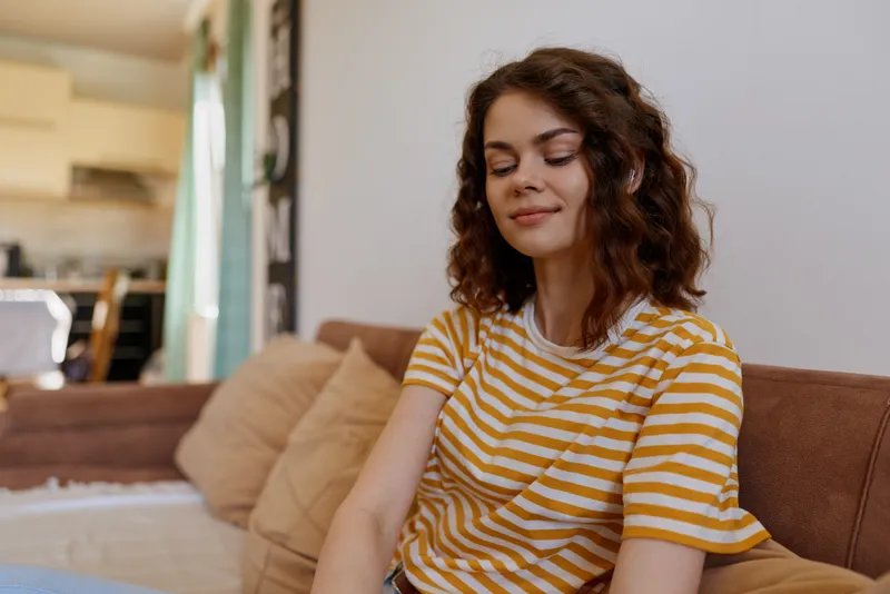A young woman with curly hair sits on a sofa at home wearing a striped yellow tshirt, relaxed and content, enjoying a calm, cozy moment in a sunlit living room surrounded by soft cushions