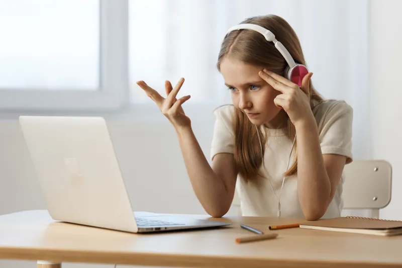Young girl expressing frustration while learning online, wearing headphones and looking at her laptop in a bright room