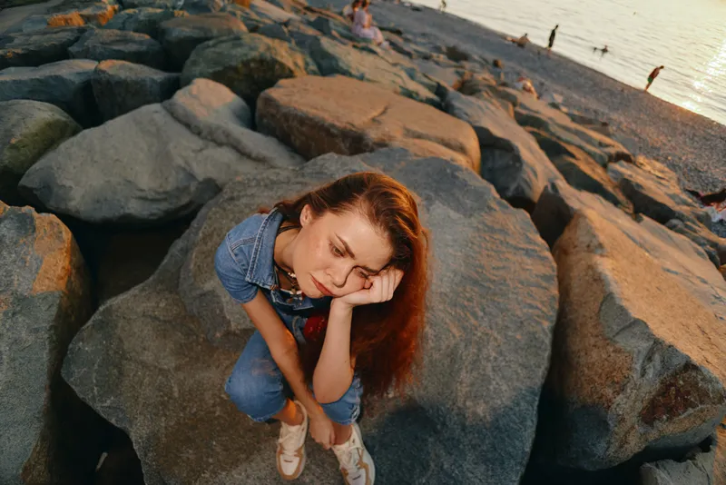 lifestyle woman in modern boho-western denim street style sitting on rocks at beach with warm film color tones, thoughtful mood, casual summer outfit