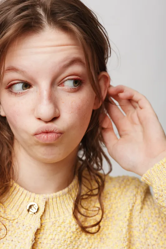 Young woman with curly hair wearing yellow sweater looks sideways with skeptical expression. Isolated on white background. Concept of doubt, curiosity, and casual fashion.