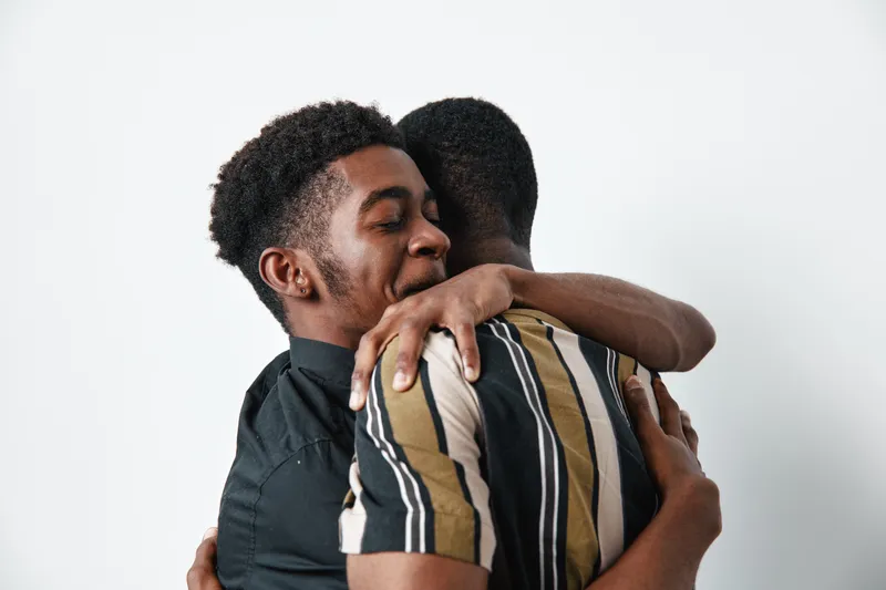 Two young black men hugging each other with eyes closed in a warm embrace. African ethnicity, dark skin, casual clothing, emotional connection, friendship and support on white background.