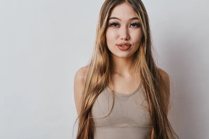 Young woman with long hair wearing sleeveless top posing against plain light background, looking at camera with slight smile and natural makeup for portrait photography.