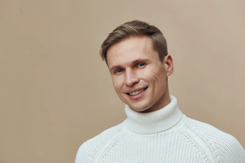 Smiling man with light brown hair wearing white turtleneck sweater looks at camera isolated on plain beige background studio portrait. People lifestyle concept, casual fashion, positive expression.