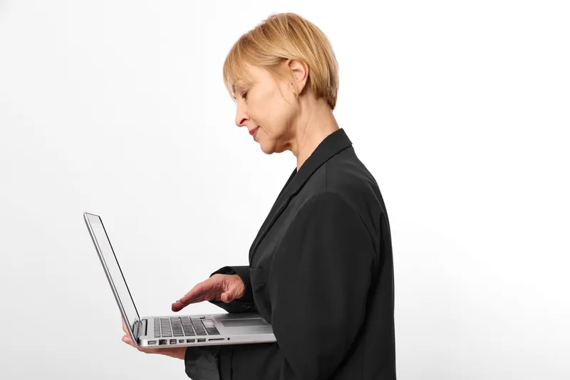 Woman typing on laptop in side profile. Senior in office setting, working on communication. Soft studio lighting shows technology and mild stress at desk during business hours.