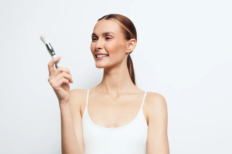 Smiling woman with clear skin holding a makeup brush, applying cosmetic product. White background, natural light, beauty and skincare concept, casual style, fresh and clean look.