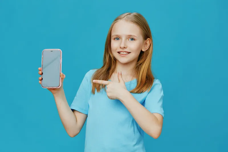 Young girl showing smartphone, smiling and pointing, against a bright blue background, wearing a casual light blue t shirt, playful and cheerful expression