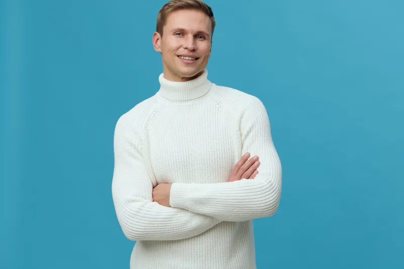 smiling young man wearing white turtleneck sweater standing arms crossed looking at camera blue background mental health and gadgets concept