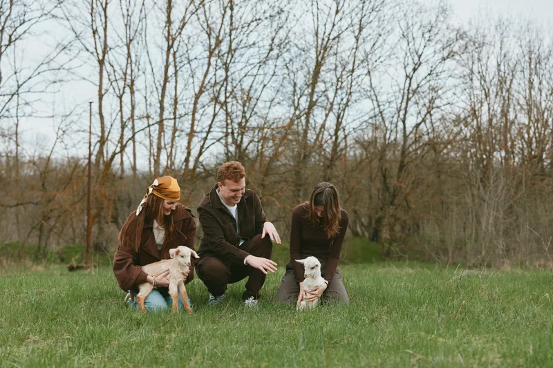 Three people in casual autumn clothing interact with two goats in a grassy park, capturing a warm lifestyle moment outdoors, highlighting family, friends, nature, and animals in a tranquil field