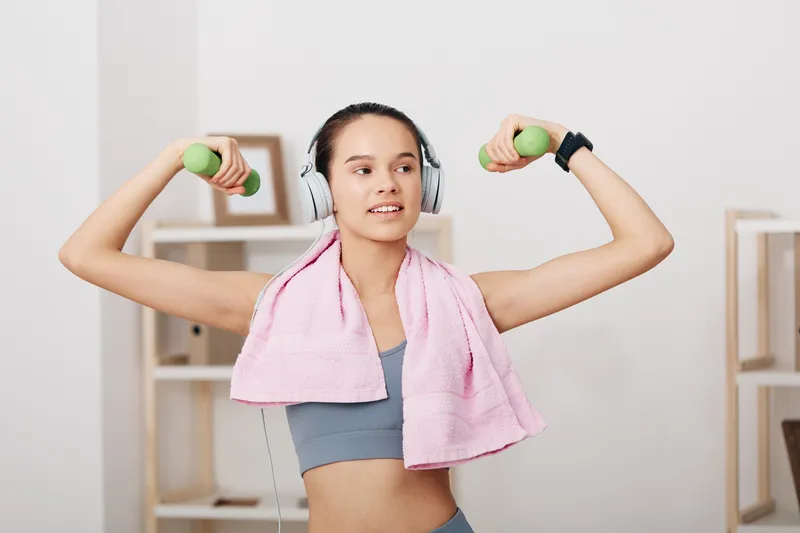 Fitness themed image of a woman in a home gym lifting green dumbbells with a pink towel over her shoulders, wearing headphones for workout motivation and a focused expression.