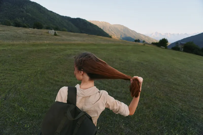 Young woman outdoors in nature, holding her long hair, casual outfit, green grass field, mountainous landscape, peaceful moment, lifestyle concept