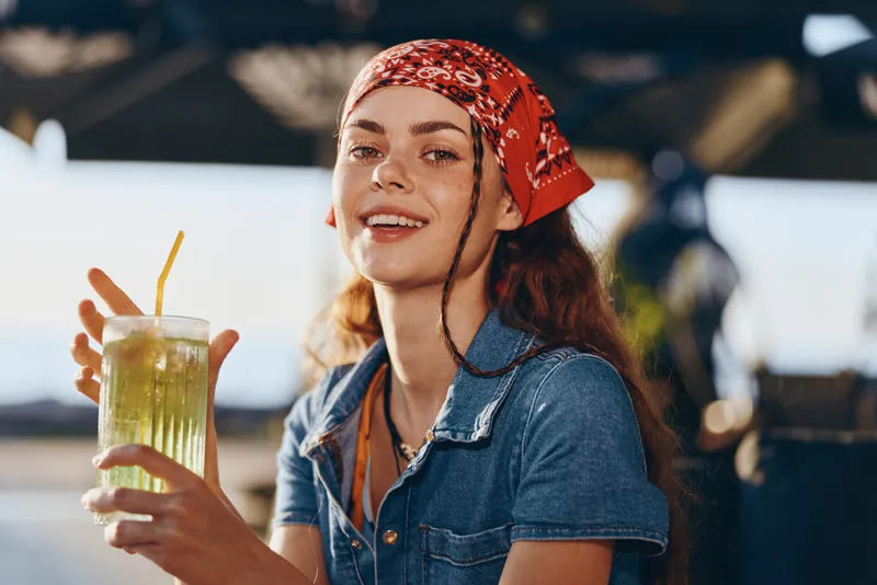 smiling woman holding refreshing drink wearing red bandana and denim shirt outdoors in sunny summer day with relaxed mood and natural light